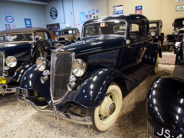 Eine Gruppe von Oldtimern in einer Garage geparkt, mit einer Wand im Hintergrund, die mit Brettern und Lampen an der Oberseite verziert ist.