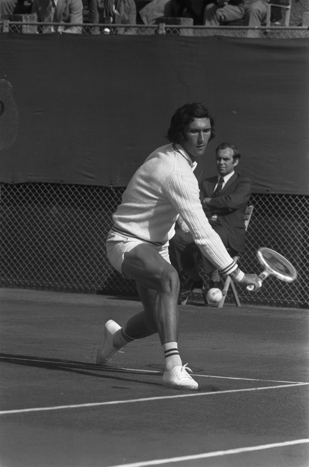 Ein Mann in sportlichem Schuhwerk schwingt einen Tennis-Sch├Ąlter gegen einen Ball, mit einem Maschendrahtzaun und Zuschauern im Hintergrund, die auf St├╝hlen sitzen.