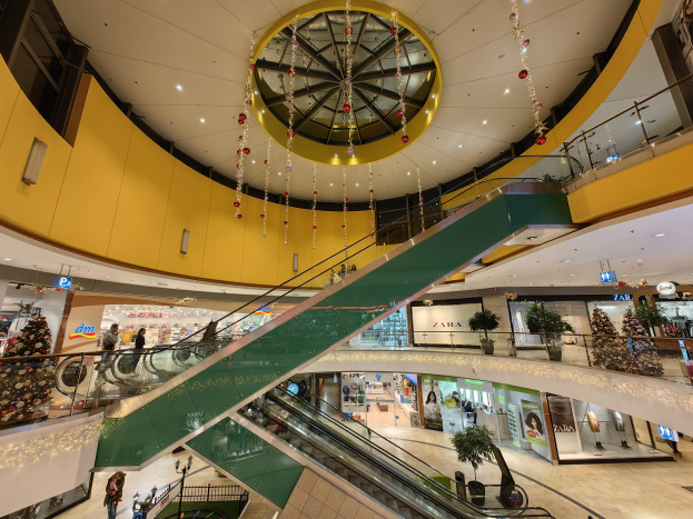 Ein belebtes Einkaufszentrum-Innere mit Rolltreppen, Gelöndern, Topfpflanzen, Geschöften, Texttafeln, Deckenlampen und hängenden Dekorationen, mit einigen Menschen.