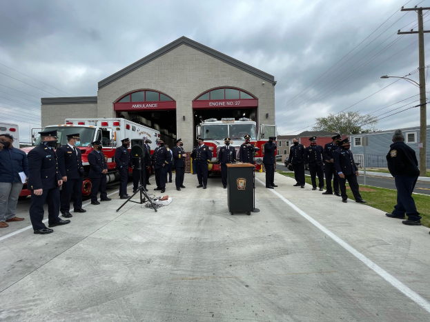 Eine Gruppe von Menschen in Mützen steht vor einem Feuerwehrauto bei einer Zeremonie für eine neue Feuerwache, mit einem Podium und Mikrofon im Vordergrund und Gebäuden, Bäumen und Strommasten im Hintergrund.
