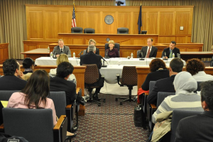 Ein Tagungsraum mit einer Gruppe von Menschen, die auf Stühlen sitzen, zwei ausländische Flaggen, ein oranger Schreibtisch mit Wasserflaschen und einem Mikrofon.