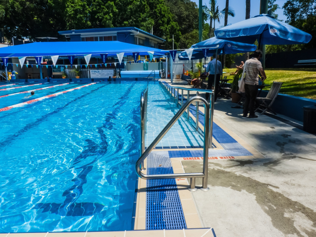 Großer Swimmingpool mit schwimmenden Menschen, umgeben von Geländern, Sonnenschirmen, Stühlen, Tischen, Pflanzen, Gras, Bäumen und einem Gebäude unter einem klaren blauen Himmel.