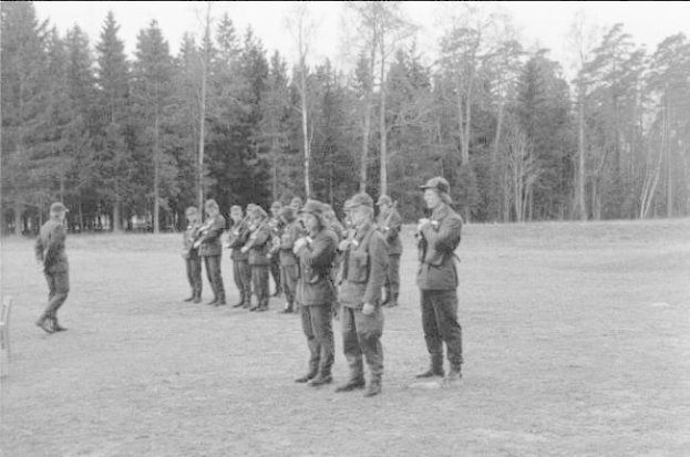 Schwarz-weißes Bild einer Gruppe von Männern in Militäruniformen mit Mützen und Gewehren, die in einem Feld mit Bäumen und einem klaren Himmel im Hintergrund stehen.