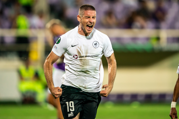 Ein Mann in weißem T-Shirt und schwarzen Shorts rennt auf einem Fußballfeld, feiert ein Tor und wird von einer Gruppe Menschen umgeben, während der Hintergrund unscharf ist.