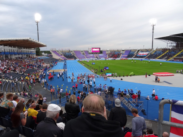 Ein großes Stadion voller Zuschauer bei einem Fußballspiel, mit Sitzplätzen und Stehplätzen, Geländern, einer Fahne, Flutlicht, einem Bildschirm, Bäumen und einem bewölkten Himmel im Hintergrund.
