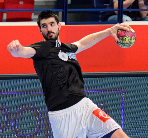 Ein Mann in schwarzem T-Shirt und weißen Shorts hält einen Ball und spielt Handball in einem Stadion mit Zuschauern im Hintergrund.