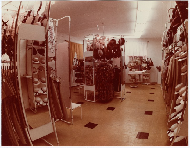 Altes Foto eines Kleiderladen-Interieurs mit Kleidung an Bügeln, Hüten auf Ständern, einem Spiegel, einem Stuhl, einem Tisch mit Gegenständen, Vorhängen und Deckenleuchten.