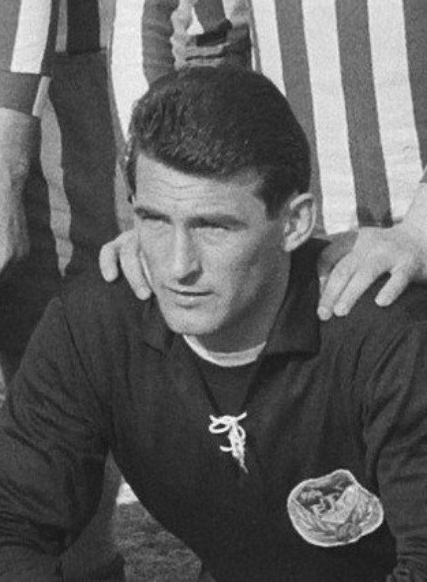 Schwarzes und weißes Foto eines Mannes, der auf einem Rasenfussballfeld sitzend ein T-Shirt und eine Kette trägt, mit zwei Personen dahinter.