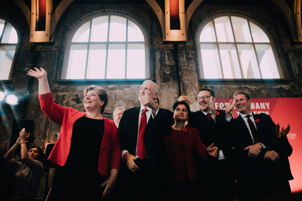 Eine Gruppe von Labour-Parteimitgliedern steht zusammen klatschend in Feierlaune vor einer Menge, mit einem Banner und beleuchteten Fenstern im Hintergrund.