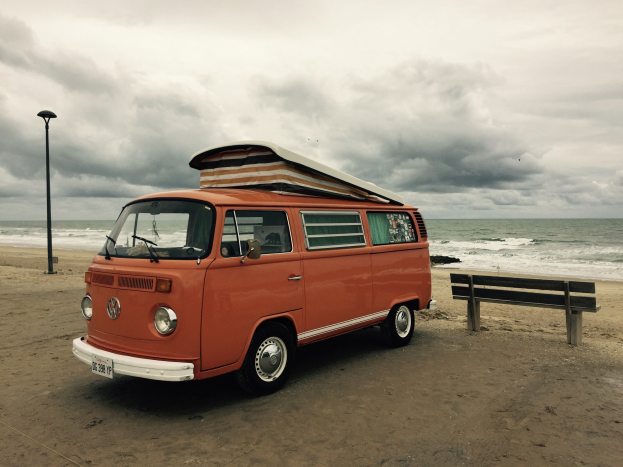 Ein oranger VW-Bus auf einem Strand neben dem Ozean geparkt, mit einer Bank und einem Laternenmast in der N├Ąhe unter einem bew├Âlktem Himmel.