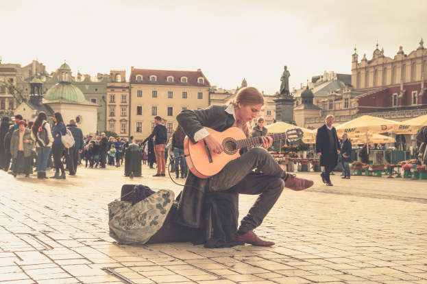 Ein Mann sitzt auf dem Boden und spielt Gitarre in einem belebten Stadtplatz, umgeben von Passanten und einer Tasche auf dem Weg, mit Gebäuden, einer Statue und einem klaren blauen Himmel im Hintergrund.