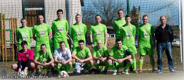 Eine Gruppe von Männern in grünen T-Shirts steht und hockt auf einem Fußballfeld mit einem Ball davor, ein Torpfosten rechts und ein Hintergrund aus Bäumen, Gebäuden, Pfählen, Fahrzeugen und dem Himmel.