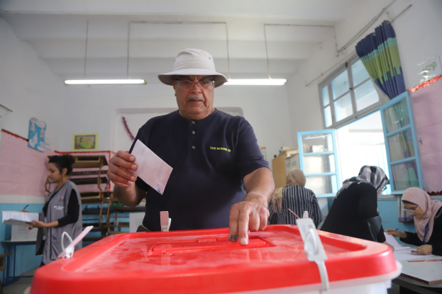 Ein Mann mit Hut stimmt bei einer Wahlurne ab, vor einer roten Wahlurne mit einem Blatt Papier in der Hand.