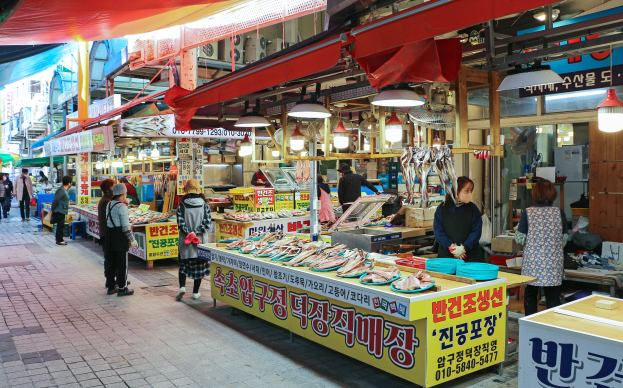 Ein belebter Straßenmarkt in Seoul, Südkorea, mit Menschen, die spazieren gehen, Verkäufern, die Artikel wie Lichter und Schilder verkaufen, und Gebäuden im Hintergrund unter einem klaren blauen Himmel.