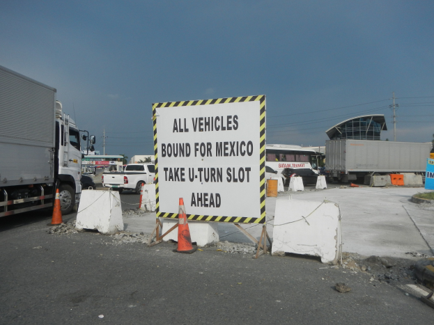 Ein Lastwagen auf einem Parkplatz neben einem Schild mit der Aufschrift "Alle Fahrzeuge nach Mexiko" geparkt, mit verstreuten Verkehrskegeln, Brettern und anderen Gegenständen im Hintergrund von Gebäuden, Bäumen, Polen und einem klaren blauen Himmel.