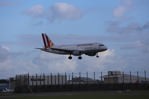 Germanwings Airbus A320-200 beim Start vom Frankfurter Flughafen mit Gras darunter, einem Zaun im Vordergrund, Gebäuden und Bäumen im Hintergrund und einer bewölkten Himmel.