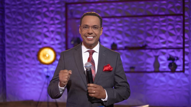 Stephen Colbert, Moderator von 'The Daily Show with Trevor Noah', steht auf einer Bühne und hält ein Mikrofon in der Hand, trägt einen Anzug, eine Krawatte und eine Brille, wobei hinter ihm ein Licht und ein Ständer zu sehen sind.