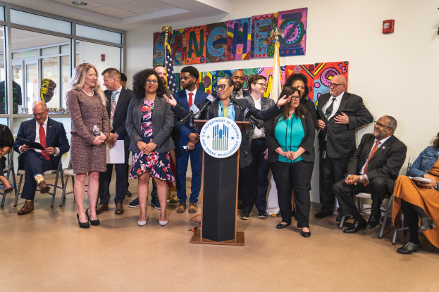 Gruppe von Menschen in einem Raum mit einem Podium, einem Mikrofon und einer Tafel mit der Aufschrift "Sancho Stadtrat tritt dem Stadtrat bei", neben Stühlen, Flaggen und einem Feuerlöscher an der Rückwand.