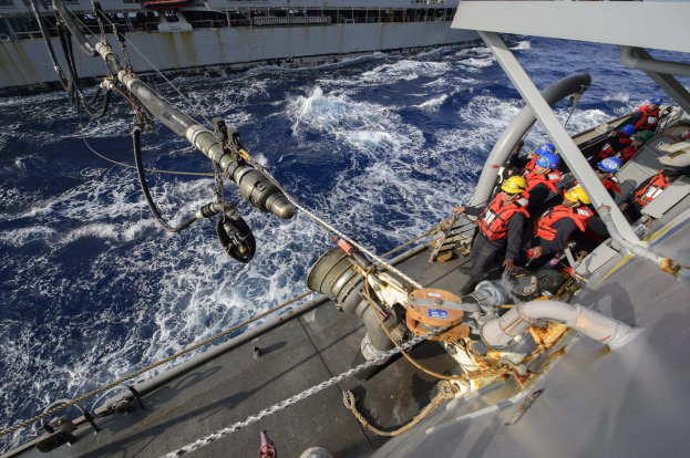 Gruppe von Menschen in Lebenswesten und Helmen auf einem Boot im Meer mit einem Schiff und einem klaren Himmel im Hintergrund.
