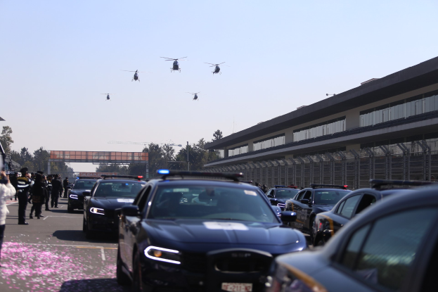 Polizeifahrzeuge vor einem Gebäude mit Menschen auf der linken Seite, Hubschrauber am Himmel und eine Brücke im Hintergrund bei klarem Himmel.