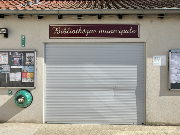 Außenansicht einer kommunalen Bibliothek mit einem 'Bibliothèque municipale'-Schild, einem zentralen Rollladen, Infotafeln, Plakaten an der linken Wand und einem beleuchteten Weg zur Einfahrt.
