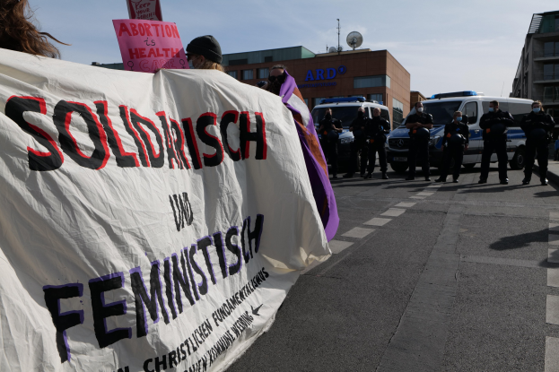 Eine Gruppe von Menschen marschiert die Straße entlang und hält ein Transparent mit der Aufschrift 'Solidarität und Feminismus', mit parkenden Fahrzeugen und Gebäuden im Hintergrund unter einem klaren blauen Himmel.
