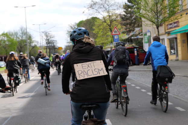 Eine Gruppe von Radfahrern, angeführt von einer Frau mit einem Schild mit der Aufschrift "Exit Racism", fährt eine von Bäumen gesäumte Straße entlang an Gebäuden und Laternen vorbei unter einem sichtbaren Himmel.