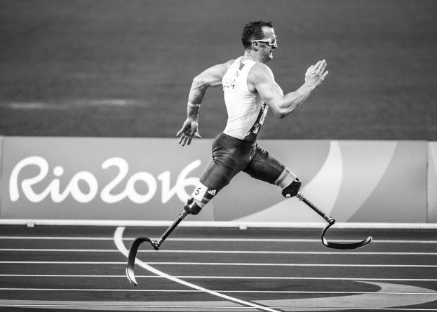 Schwarzes und weißes Foto eines Mannes mit einer Prothese, der auf einer Laufbahn läuft, mit einer Tafel im Hintergrund, auf der "Rio 2016 Paralympische Spiele" steht.