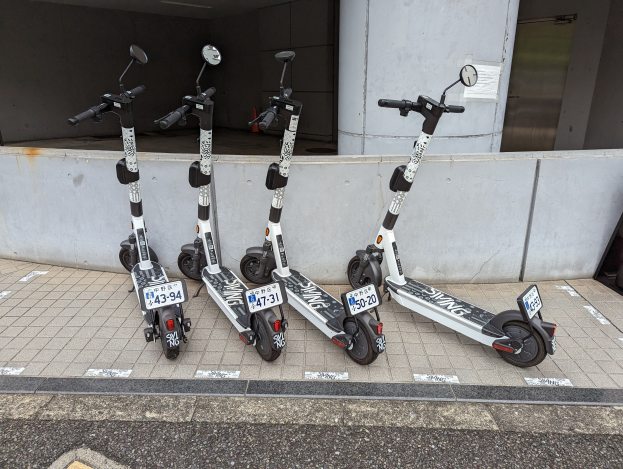 Drei schwarze und weiße Elektro-Scooter mit Nummern und Text sind nebeneinander auf einem Gehweg vor einem Gebäude mit einer Wand und einer Glastür im Hintergrund geparkt.
