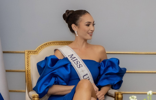 Frau in einem blauen Kleid mit einer "Miss Universe 2019" Schärpe, die auf einem Stuhl sitzt und von Blumen umgeben ist, mit einem Lächeln vor einer Wand.