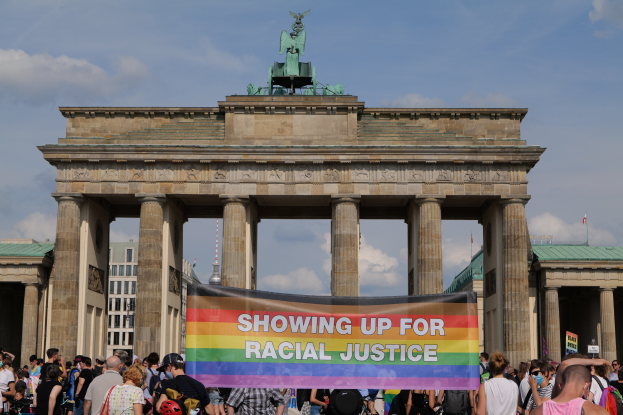 Eine Gruppe von Menschen hält eine "Racial Justice"-Schriftzug vor dem Brandenburger Tor in Berlin, mit den Säulen und der Statue des Tors im Hintergrund und Gebäuden und bewölktem Himmel.