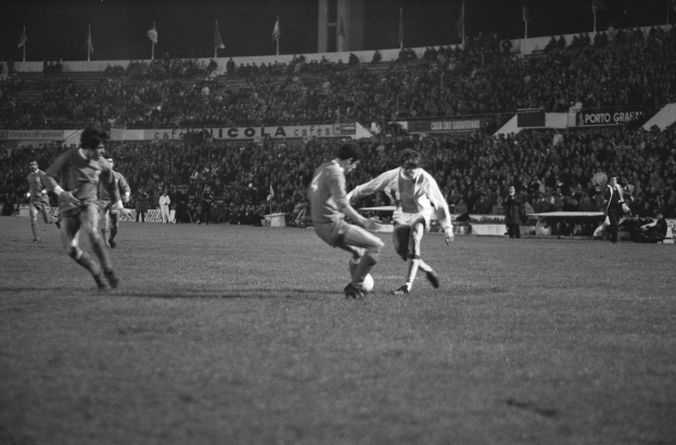 Schwarzes und weißes Bild von Männern, die Fußball auf einem Feld in einem Stadion spielen, mit Zuschauern auf den Tribünen und Fahnen im Hintergrund.