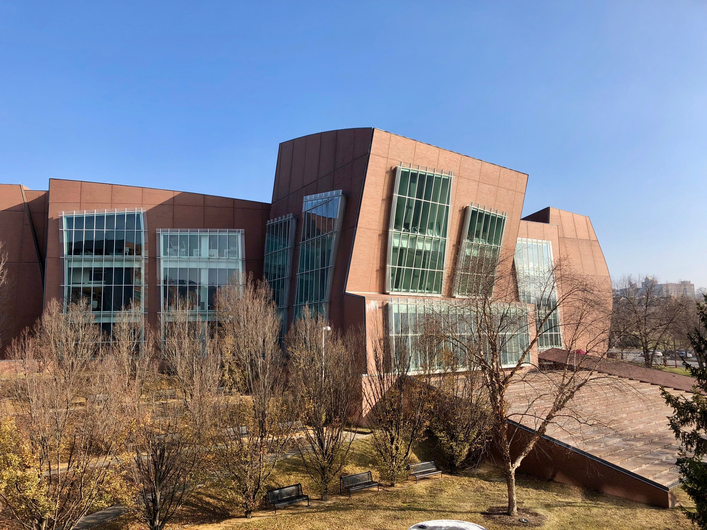 Großes modernes Gebäude mit vielen Fenstern, umgeben von Bäumen, Bänken, Gras und einem Laternenmast, mit anderen Gebäuden und einem klaren blauen Himmel im Hintergrund, als der neue Campus der University of Illinois at Chicago identifiziert.