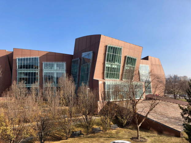 Großes modernes Gebäude mit vielen Fenstern, umgeben von Bäumen, Bänken, Gras und einem Laternenmast, mit anderen Gebäuden und einem klaren blauen Himmel im Hintergrund, als der neue Campus der University of Illinois at Chicago identifiziert.
