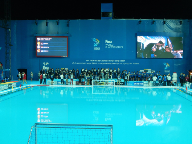 Gruppe von Menschen um ein Hallenbad mit Netz am Boden herumstehend, mit einem Bildschirm im Hintergrund, der "FINA World Championships 2015 Kazan" anzeigt.