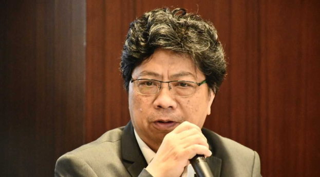 Ein Mann im Anzug mit einem Mikrofon, der lebhaft gegen eine hölzerne Wand spricht.