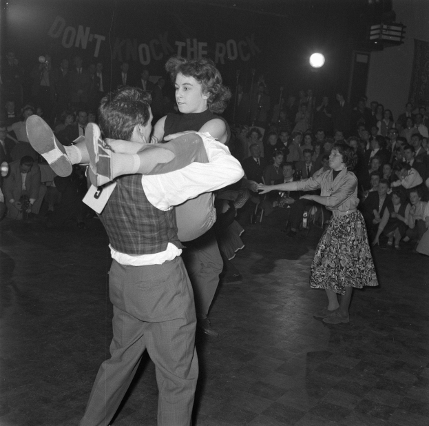 Ein Mann und eine Frau tanzen gemeinsam vor einem Publikum, das von einem Lichtstrahl beleuchtet wird, in einem Schwarz-Weiß-Bild.
