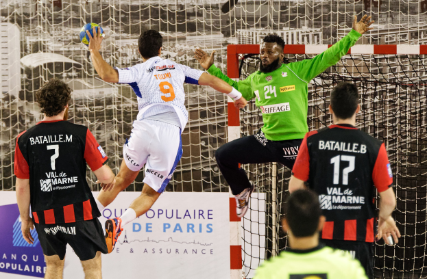 Eine Gruppe von Männern spielt Handball, einer springt und hält den Ball, vor einem Torpfosten und einer Fahne mit der Aufschrift "Ligue 1 - Bordeaux vs Marseille".