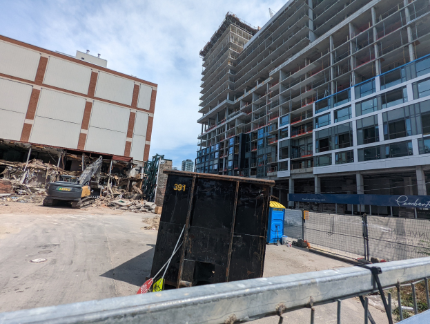 Ein großes Gebäude im Bau mit Schutt davor, umgeben von einem Metallzaun, einem Container, Pflanzen, Bäumen, Pfählen, einem Schild, Fahrzeugen, Steinen und einem bewölkten Himmel.