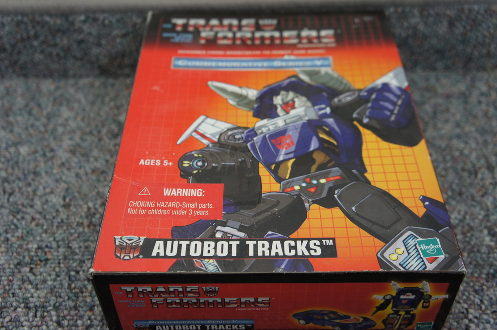 Ein roter, oranger und blauer Kasten mit einem Roboter und dem Text "AUTOBOT TRACKS" darauf, vor einem unscharfen schwarzen Hintergrund.