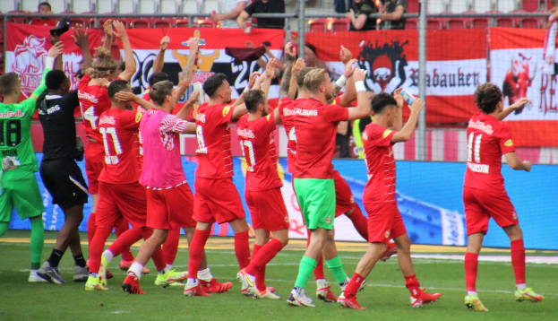 Gruppe von Fußballspielern, die ein Tor feiern, auf einem Feld mit Flaschen haltenden oder klatschenden Spielern, Bannern, einem Zaun, Stühlen und einer Dachkonstruktion im Hintergrund.