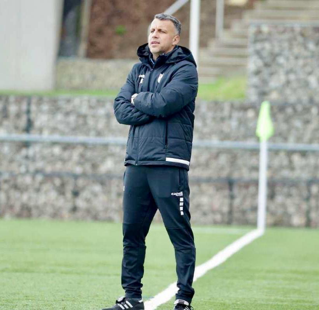 Mann auf Fußballfeld mit verschränkten Armen in schwarzer Jacke und Hose, im Hintergrund ein Zaun, Treppe und Wand