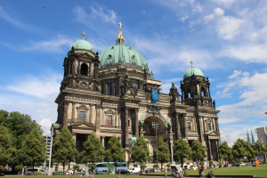 Der Berliner Dom in Berlin, Deutschland, ein großes Gebäude mit Fenstern, Säulen, Bögen und Statuen, gekrönt von einem Kreuz, mit Menschen auf der Wiese davor, Fahrzeugen, Bäumen, Pfählen, Flaggen und einem bewölkten Himmel im Hintergrund.