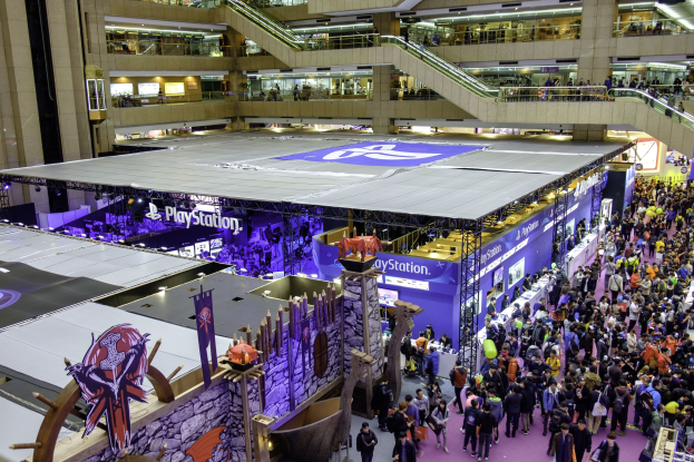 Eine große Menschenmenge in einem großen Gebäude mit Rolltreppen, Geländern, beleuchteten Schildern, Bannern und anderen Convention-Ausstellungsstücken, mit einem Wasserzeichen, das darauf hinweist, dass es die größte Gaming-Convention der Welt ist.