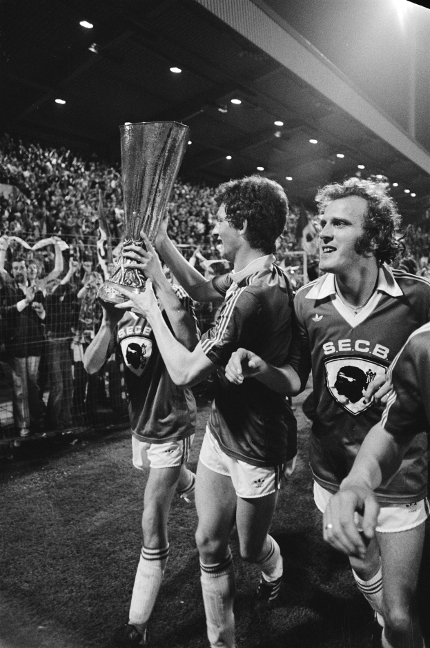 Schwarzes und weißes Bild von Fußballspielern, die einen Pokal auf einem Feld halten und ihren Sieg im FA-Cup-Finale feiern, während Zuschauer hinter einem Zaun zusehen.