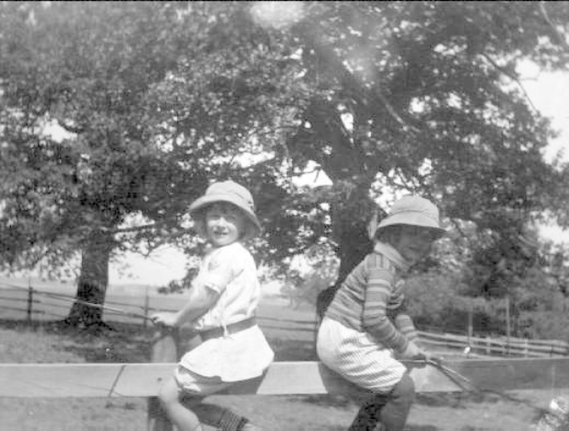 Zwei junge Jungs in Hüten mit Baseballschlägern in einem grasigen Park mit Bäumen und einem Zaun im Hintergrund, Schwarz-Weiß-Foto.
