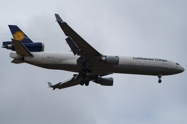 Ein Lufthansa Cargo Boeing 777-300ER im Flug, mit ausgefahrenen Flügeln und sichtbaren Triebwerken, mit 'Lufthansa Cargo' Schriftzug auf dem Rumpf vor einem bewölkten Himmel.