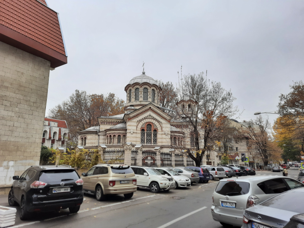 Eine Straßenszene mit parkenden Autos, Gebäuden, Bäumen, Laternenpfählen und einem bewölkten Himmel, mit einem Kraftfahrzeug-Kennzeichen im Vordergrund.