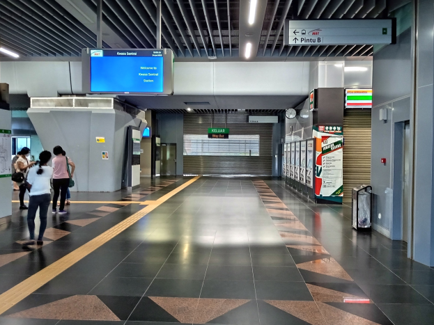 Menschen gehen durch einen Bahnhof mit einem Kofferwagen auf der rechten Seite, Schilder mit Text, eine Uhr an der Wand und Deckenbeleuchtung.