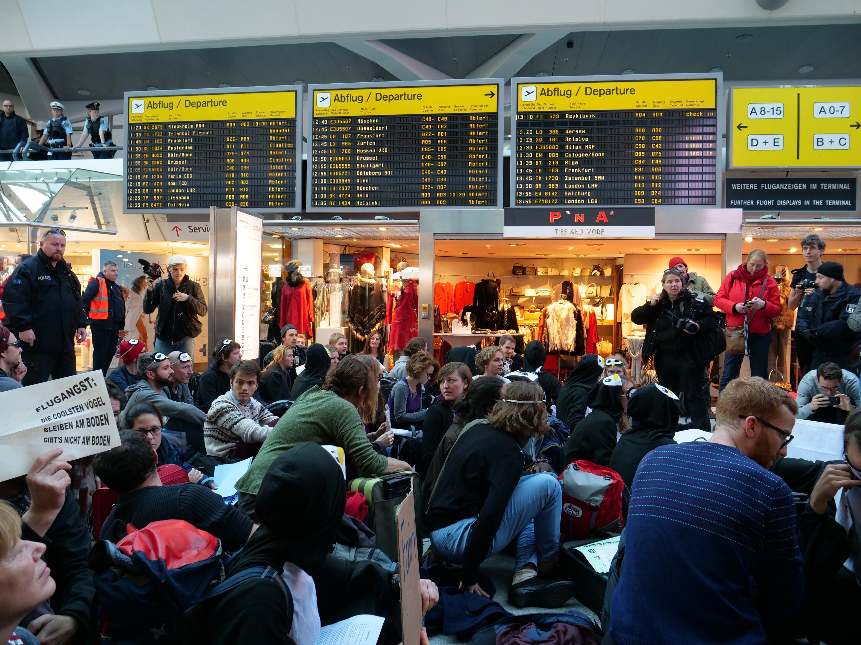 Eine große Gruppe von Menschen sitzt und steht in einem Flughafen während einer Protestaktion, mit Schildern, Puppen in Kleidern und Deckenleuchten im Hintergrund.